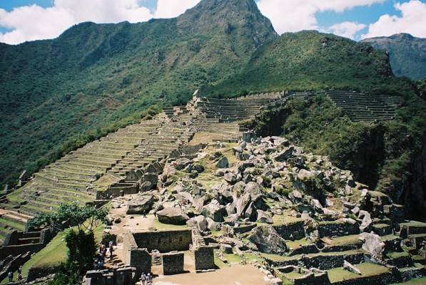 There are terraces covered in grass going down the side of a mountain, there is also evidence of the ruins of a building and you can see large rocks on the side of the mountain too.  The whole area is in the mountains and what is not excavated is covered in trees