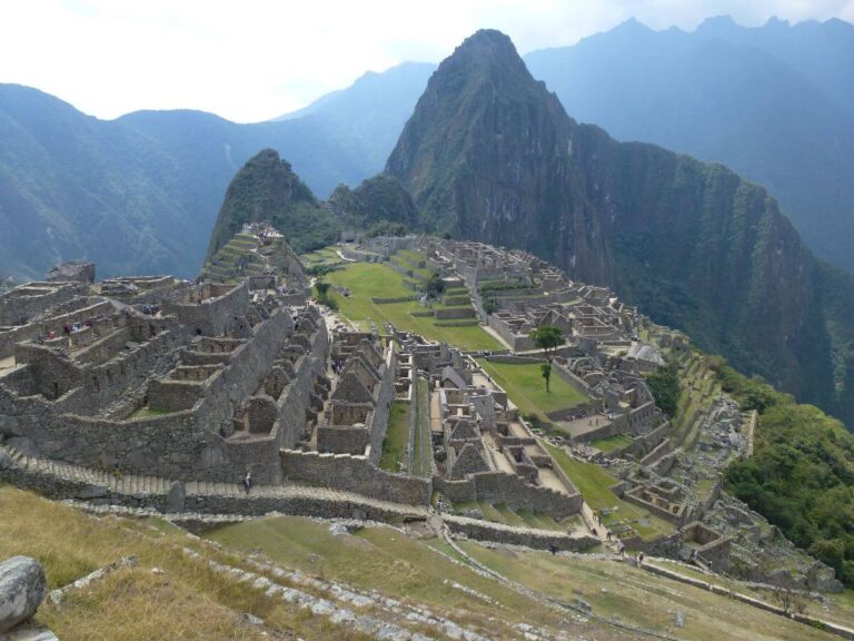 a view from high up of some ruins on the top of a mountain, there is evidence of many buildings and terraces, the area is covered in grass and you can see mountains covered in trees in the background