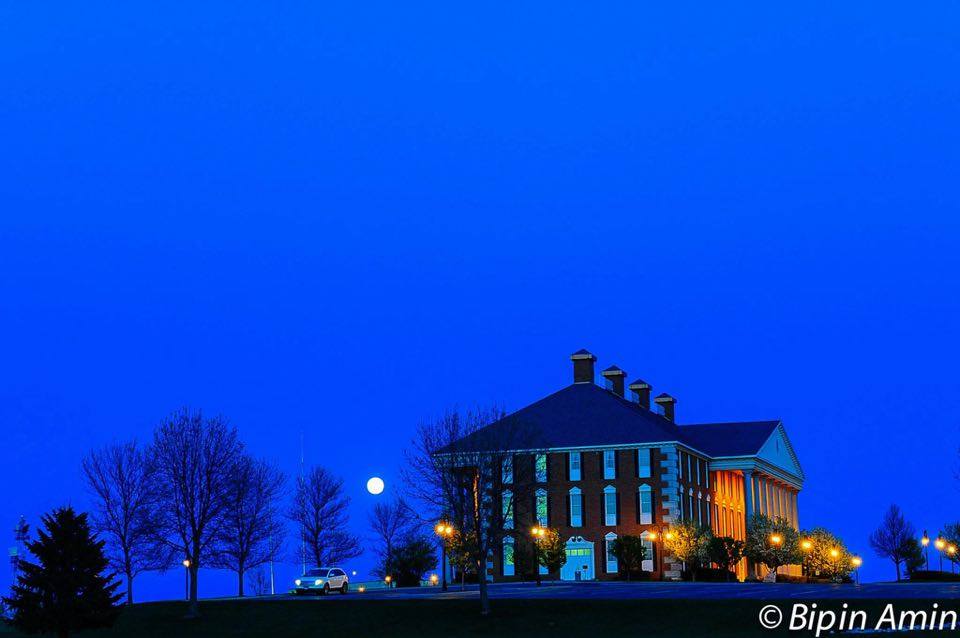Redlin Art Center Watertown, South Dakota. A stately brick building illuminated by warm lights at dusk, set against a deep blue evening sky, with bare trees, glowing streetlamps, and a full moon rising near the horizon.
