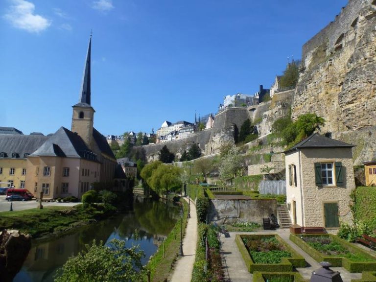 A scenic view of Luxembourg City’s Grund district on a sunny day, featuring a narrow canal bordered by manicured gardens and lush greenery. To the left stands a tall church with a sharp spire, reflected in the calm water below. On the right, historic stone fortification walls rise steeply, topped with traditional European buildings.