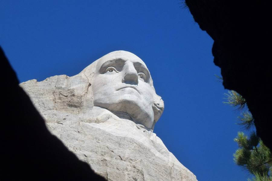 a close up image of a mans face carved into the rock from below, you can see the details of his eyes and nose and it is a beautiful sunny day