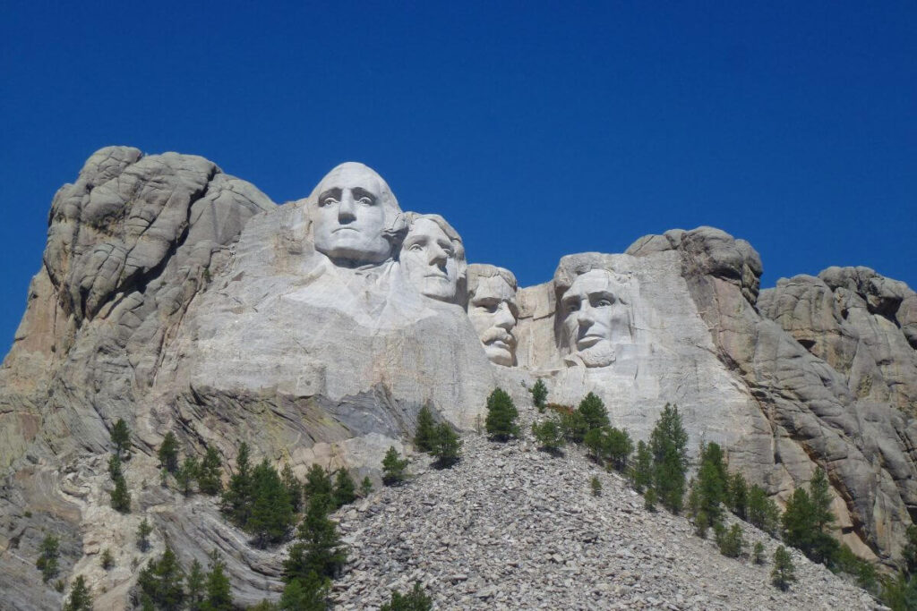 Close-up view of Mount Rushmore in South Dakota, showing the massive granite faces of four U.S. presidents carved into a rocky mountainside, with scattered pine trees on the slope below and a clear deep blue sky overhead.