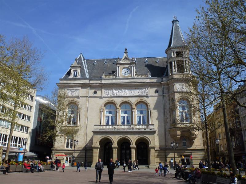 The City Palace (Cercle Cité) at Place d'Armes in Luxembourg City, a grand historic building with ornate stonework, arched windows, and a central clock above the entrance. The façade features decorative carvings and a turret on the right side. In the foreground, people stroll and relax in the sunlit square surrounded by trees and nearby cafés, creating a vibrant and inviting city atmosphere.