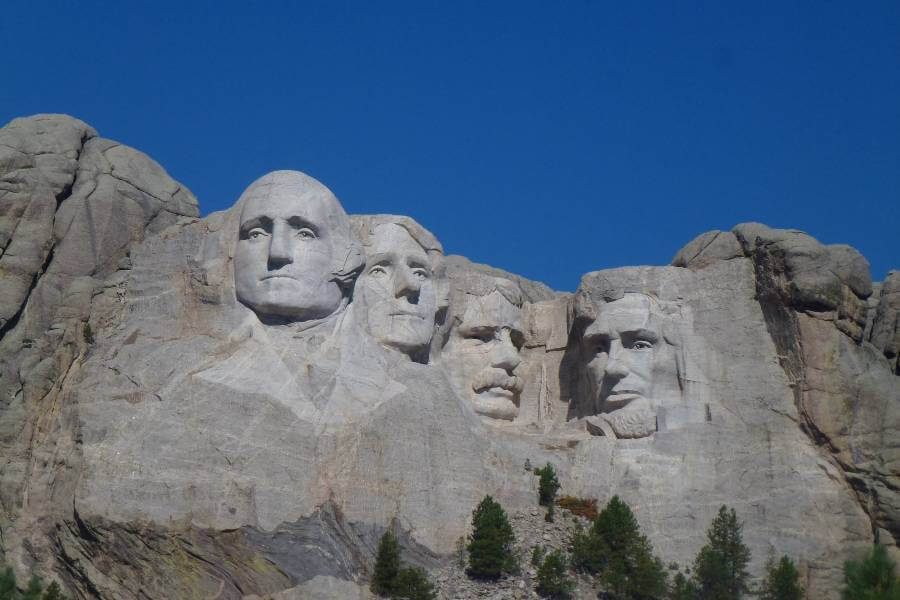4 faces carved into the side of a smooth mountain, you can see the details of the eyes, hair, noses and lips and one has a moustache, before them you can see a few trees on a beautiful sunny day