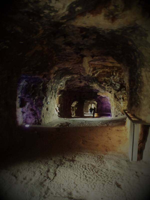 Dimly lit interior of the Bock Casemates tunnels in Luxembourg City. The image shows a series of stone passageways with arched ceilings, carved into the rock. A soft glow of purple and white lighting illuminates parts of the tunnel, adding atmosphere to the ancient space. Two people are visible walking in the distance, giving a sense of scale to the cavernous corridors. The foreground features a sandy, textured floor and a wooden railing on the right side.