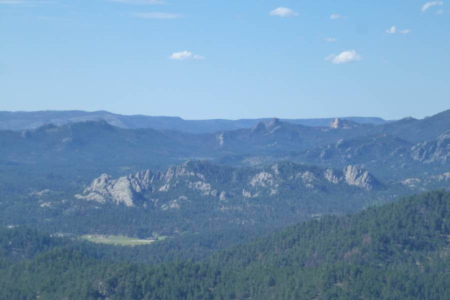 Wide view of the Black Hills in South Dakota, showing rolling, forested hills dotted with rocky granite outcrops and distant ridgelines fading into the horizon beneath a pale blue sky