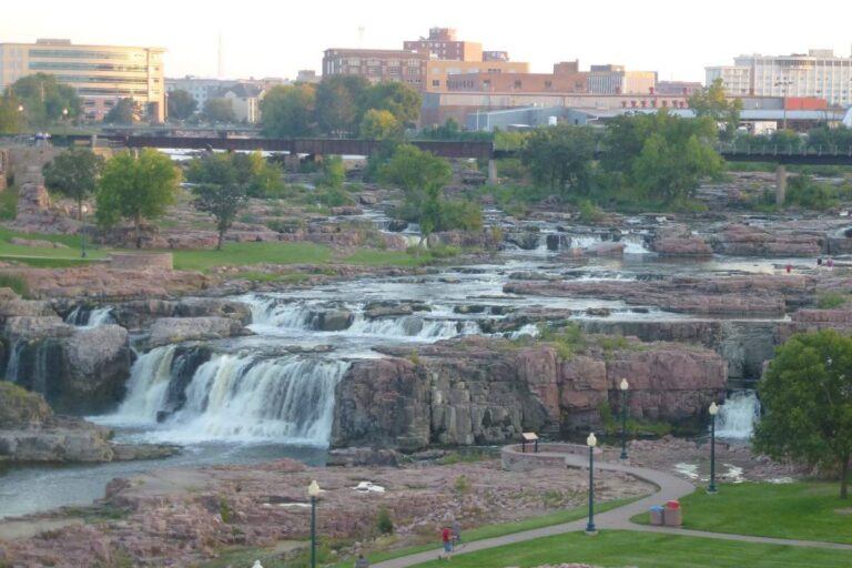 Scenic view of Sioux Falls Park in South Dakota, featuring cascading waterfalls flowing over rugged pink quartzite rock formations. A winding path with streetlamps and green grassy areas surrounds the falls, while trees add touches of greenery. In the background, a bridge and modern city buildings rise against the sky, blending natural beauty with an urban skyline.