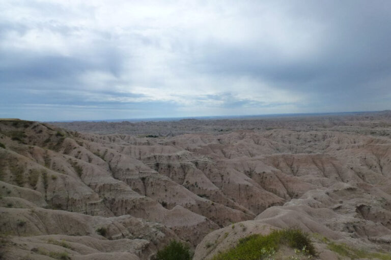 Expansive view of the rugged landscape at Hay Butte Overlook in Badlands National Park, South Dakota. The scene showcases layered rock formations and deep eroded canyons in soft pink and beige tones stretching toward the horizon under a cloudy blue-gray sky.