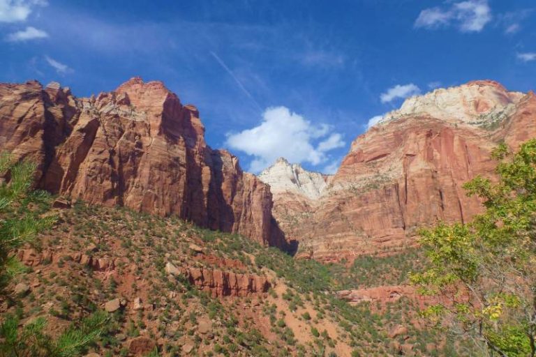 A striking landscape view of Zion National Park featuring towering red and cream sandstone cliffs rising sharply against a deep blue sky with scattered white clouds. The rocky slopes are dotted with green desert vegetation, and sunlight highlights the rich layers and textures of the canyon walls, creating a dramatic and colorful desert scene.