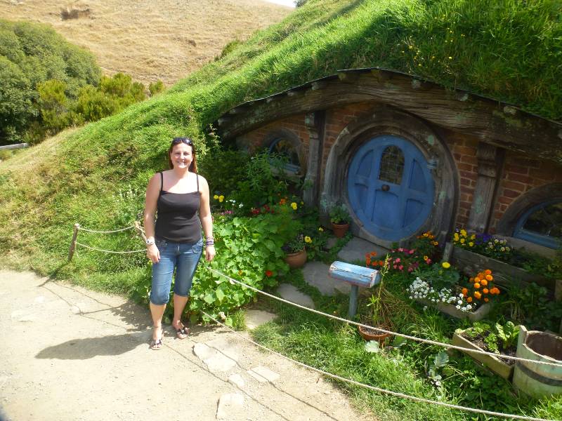 ilive4travel stood outside a hobbit hole with a blue door and lots of flowers in the garden and lots of green grass