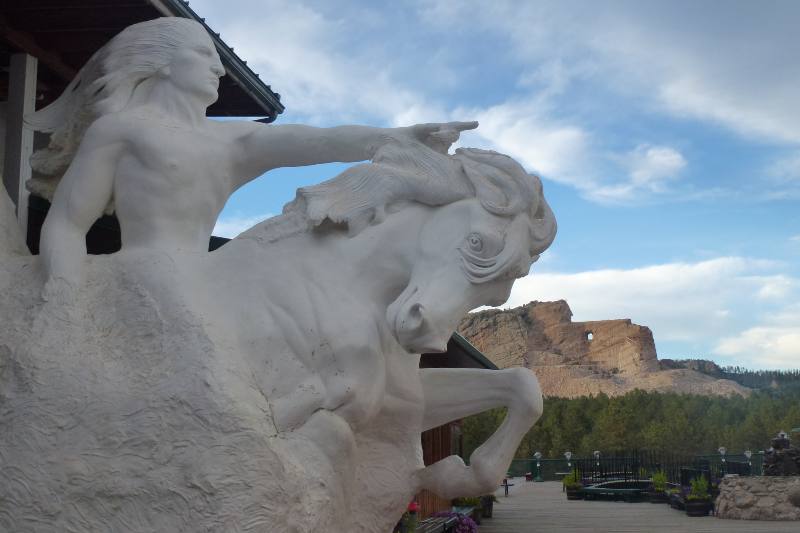 a model of crazy horse on top of his horse with the mountain carving in the background.  Crazy horse is riding his horse, he has long hair and is pointing his first finger on his left hand