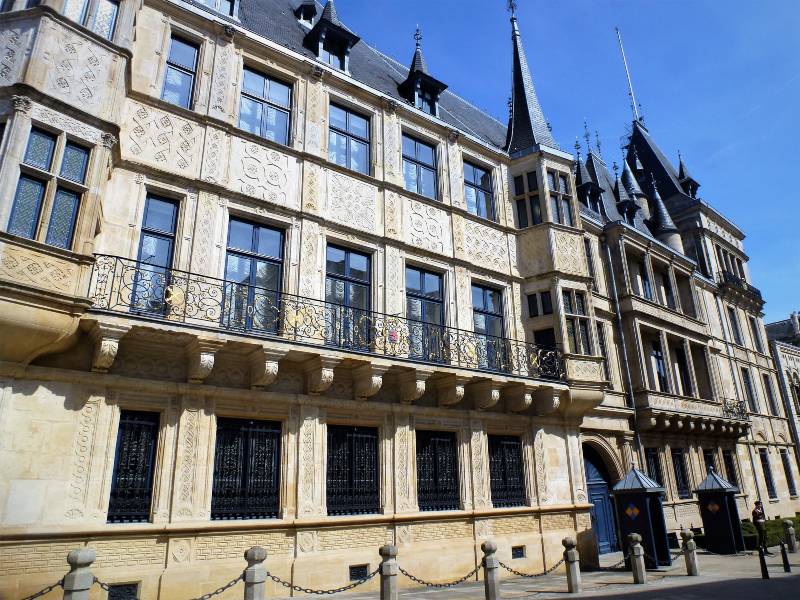 The outside of the Grand Ducal palace in Luxembourg, it is 3 storeys high with some turrets on the roof. There are railings outside the building to stop people getting too close and a guard, guarding the entrance