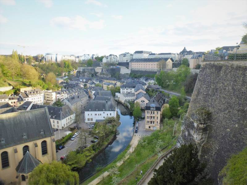 a view down into a valley with a river snaking through the middle of it, with buildings on either side of the river and a large wall to the right, with buildings on top of the wall