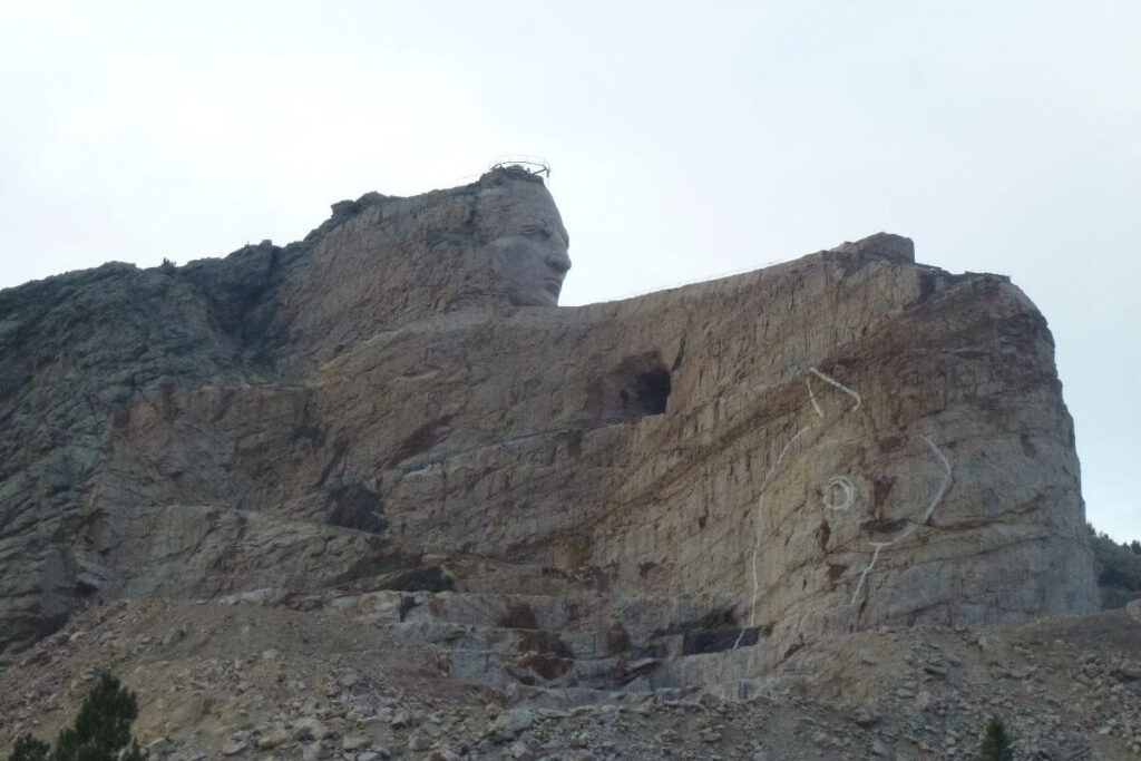 a mountain at Crazy Horse Memorial South Dakota carved with a side on view of a face, there is also other working going on to the mountain to the right of the face which is flat and there is an image of a horse head drawn onto the rock of the mountain