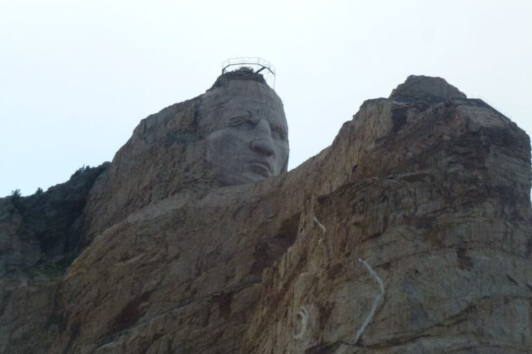 Close-up view of the Crazy Horse Memorial in South Dakota, showing the detailed face of Crazy Horse carved into a granite mountain. The sculpture is still under construction, with scaffolding visible on the top of the head. The surrounding rock remains mostly uncarved, highlighting the ongoing nature of the monumental project. The sky is overcast, casting a muted light over the scene.