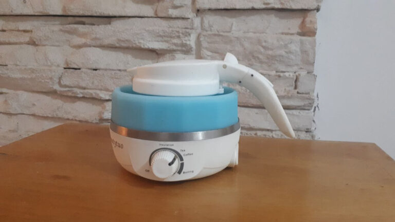 a white and blue travel kettle that has been pushed down so that it is at its smallest, sat on a wooden table with a stone wall behind it