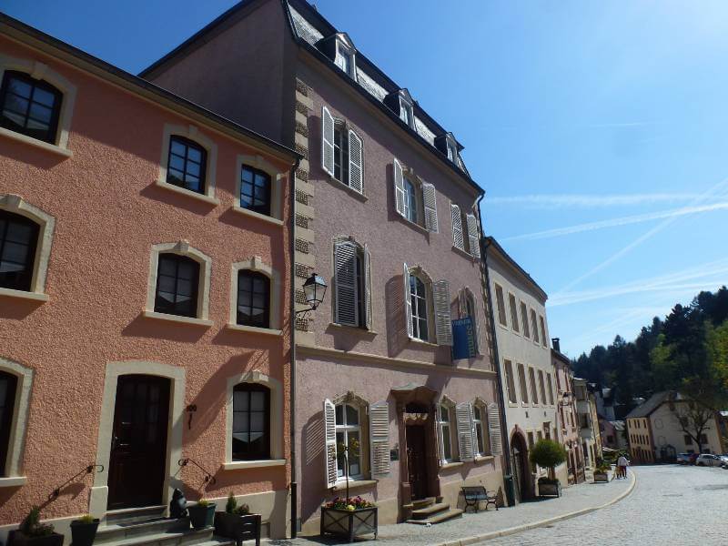 a road through a town with lots of buildings on the side of it, in orange, lilac, grey, with planters outside and one of the buildings has white shutters on the windows, in the distance you can see trees on a hill