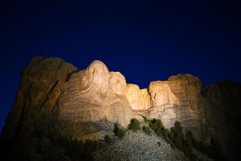 Mount Rushmore National Memorial illuminated at night during the lighting ceremony, with the carved faces of George Washington, Thomas Jefferson, Theodore Roosevelt, and Abraham Lincoln glowing against a dark blue sky. The surrounding granite cliffs and pine trees are dimly visible in the foreground.
