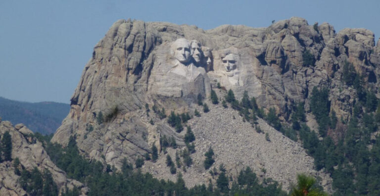 In the distance Mount Rushmore National Memorial under a blue sky, featuring the carved faces of four U.S. presidents, George Washington, Thomas Jefferson, Theodore Roosevelt, and Abraham Lincoln, set into a granite mountain surrounded by pine-covered slopes and a rocky debris field at the base.