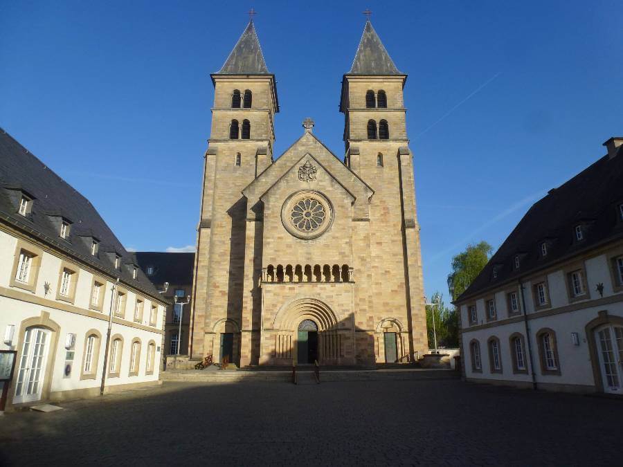 A photo of the Basilica of Saint Willibrord in Echternach, Luxembourg, showing its symmetrical twin towers, arched entrance, and rose window. The church is framed by two white buildings with slate roofs on either side under a clear blue sky.