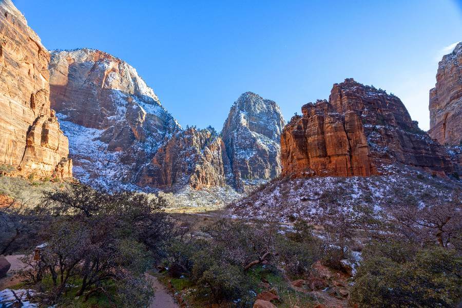a view from a path on the valley floor looking up at the orange cliffs surrounding the valley, there is a light covering of snow on the ground and a blue sky