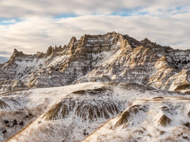 Snow-covered rock formations and rugged ridges in Badlands National Park, South Dakota, with layered cliffs and rolling hills dusted in winter snow beneath a cloudy sky.