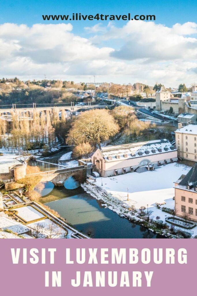 Pinterest pin showing a winter view of Luxembourg City with light snow covering the rooftops, bridges, and riverbanks. The image features the scenic Alzette River and the historic stone bridge surrounded by pastel buildings under a bright blue sky. Text on the pin reads “Visit Luxembourg in January”