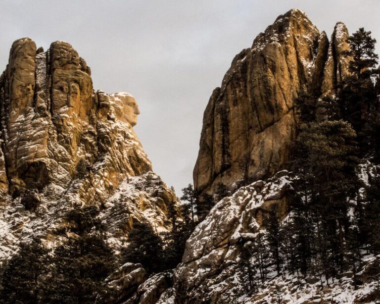 Snow-dusted granite cliffs at Mount Rushmore with George Washington’s carved face emerging prominently from the rock on the left side. The rugged golden-brown peaks frame the sculpture dramatically, while dark green pine trees cover the lower slopes beneath a cloudy gray winter sky.
