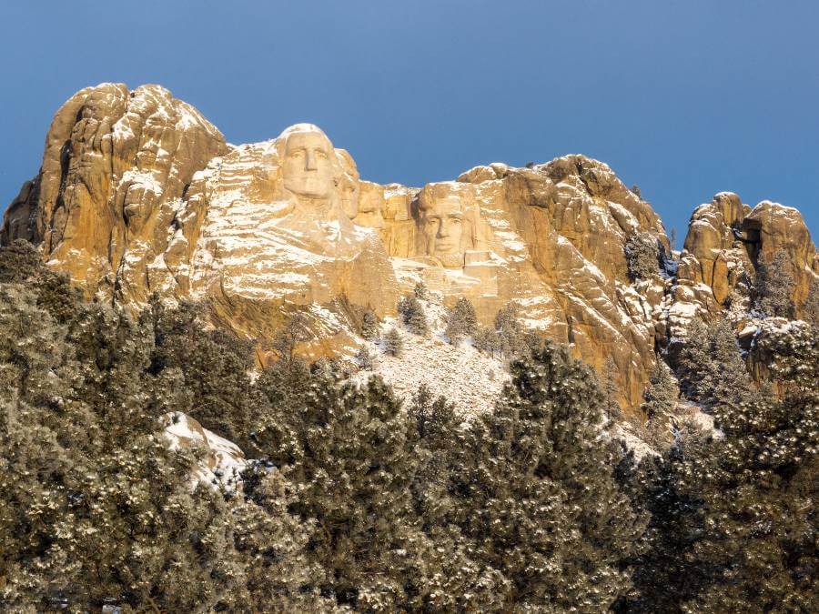 Mount Rushmore National Memorial in winter, with the carved faces of George Washington, Thomas Jefferson, Theodore Roosevelt and Abraham Lincoln partly covered in snow, surrounded by rugged granite cliffs. Evergreen trees dusted with snow fill the foreground beneath a clear blue sky.