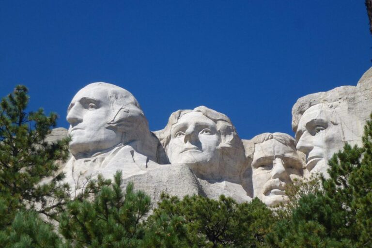 Mount Rushmore National Memorial in South Dakota, featuring the carved granite faces of George Washington, Thomas Jefferson, Theodore Roosevelt, and Abraham Lincoln, framed by green pine trees under a clear blue sky.
