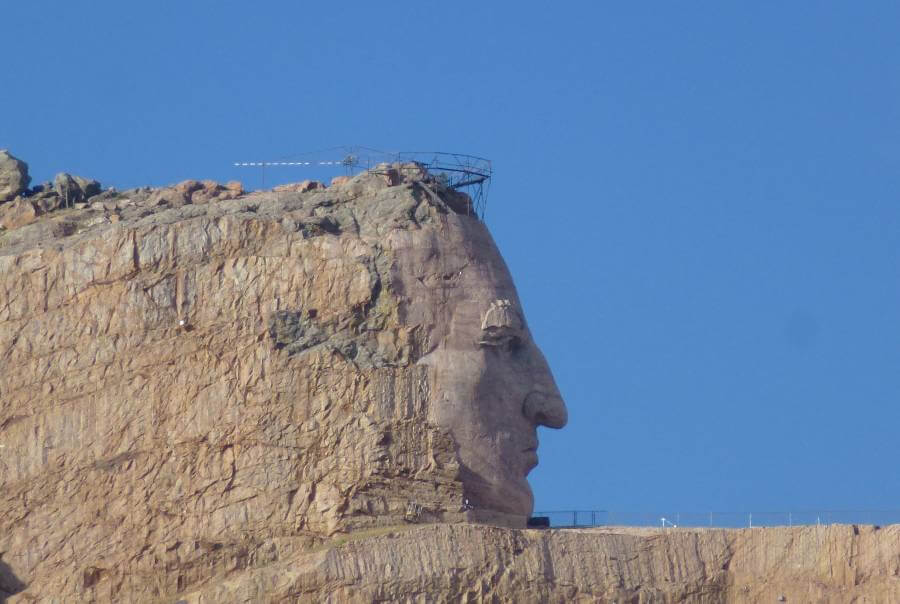 Close-up view of the Crazy Horse Memorial sculpture in South Dakota, showing the detailed carving of Crazy Horse’s face emerging from the mountain. The rock is a warm reddish-brown, and metal scaffolding is visible at the top where construction continues. The sky is clear and bright blue, highlighting the scale and unfinished state of the monument.
