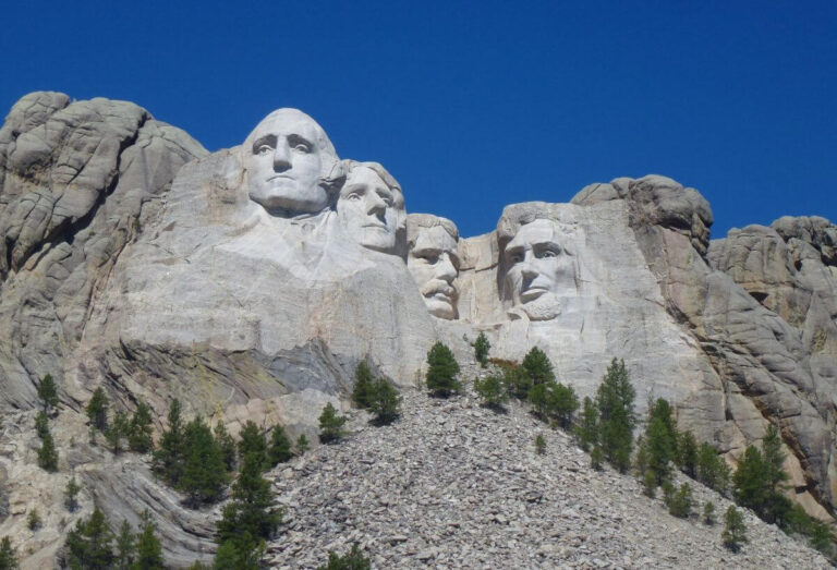 A clear daytime image of Mount Rushmore National Memorial in South Dakota, featuring the massive granite carvings of four U.S. Presidents, George Washington, Thomas Jefferson, Theodore Roosevelt, and Abraham Lincoln, set against a deep blue sky, with pine trees visible at the base of the mountain.