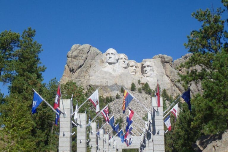 View of Mount Rushmore National Memorial with the carved faces of four U.S. presidents in the background, seen from the Avenue of Flags lined with state and territorial flags leading toward the monument under a clear blue sky.