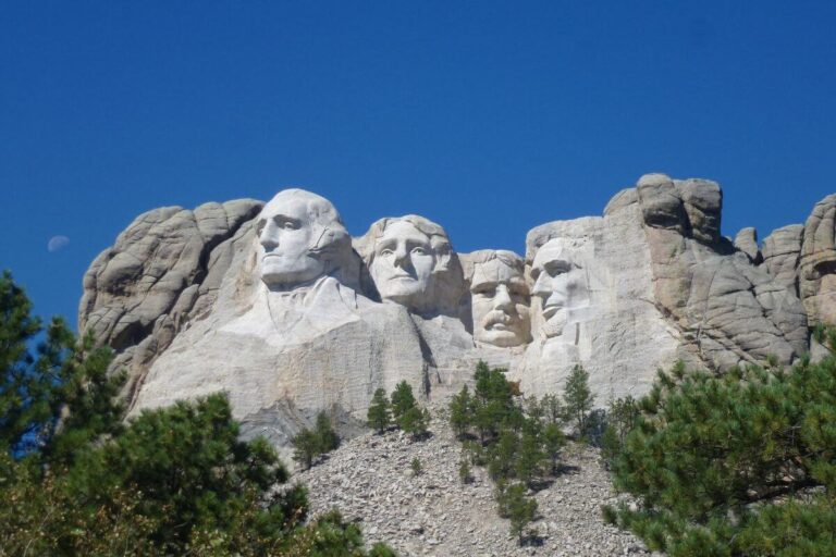 Mount Rushmore National Memorial under a bright blue sky, featuring the carved faces of four U.S. presidents, George Washington, Thomas Jefferson, Theodore Roosevelt, and Abraham Lincoln, set into a granite mountain surrounded by pine-covered slopes and a rocky debris field at the base.