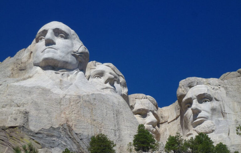 Close-up view of Mount Rushmore National Memorial under a clear blue sky, featuring the massive granite carvings of four U.S. presidents, George Washington, Thomas Jefferson, Theodore Roosevelt, and Abraham Lincoln. The white stone faces stand out against the vivid sky, with green pine trees scattered at the base of the monument.