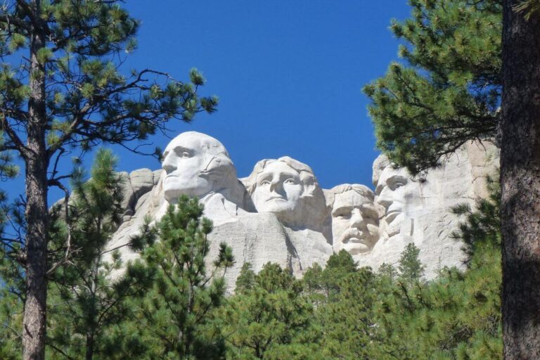 Mount Rushmore National Memorial in South Dakota, featuring the carved granite faces of George Washington, Thomas Jefferson, Theodore Roosevelt, and Abraham Lincoln, framed by green pine trees under a clear blue sky.