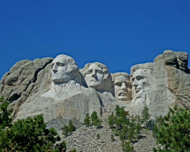 An image of Mount Rushmore National Memorial in South Dakota, featuring the massive granite carvings of four U.S. Presidents, George Washington, Thomas Jefferson, Theodore Roosevelt, and Abraham Lincoln, set against a deep blue sky, with pine trees visible at the base of the mountain.