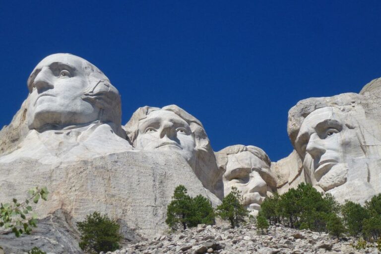 Close-up view of Mount Rushmore National Memorial under a clear blue sky, featuring the massive granite carvings of four U.S. presidents, George Washington, Thomas Jefferson, Theodore Roosevelt, and Abraham Lincoln. The white stone faces stand out against the vivid sky, with green pine trees scattered at the base of the monument.