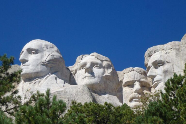 Mount Rushmore National Memorial in South Dakota, featuring the carved granite faces of George Washington, Thomas Jefferson, Theodore Roosevelt, and Abraham Lincoln, framed by green pine trees under a clear blue sky.