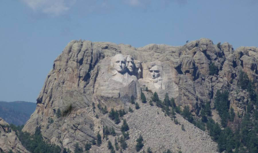 Distant view of Mount Rushmore in South Dakota, with the carved faces of four U.S. presidents, George Washington, Thomas Jefferson, Theodore Roosevelt, and Abraham Lincoln, set against rugged granite cliffs. There are green pine trees in the foreground under the monument which is under a bright blue sky with scattered white clouds.