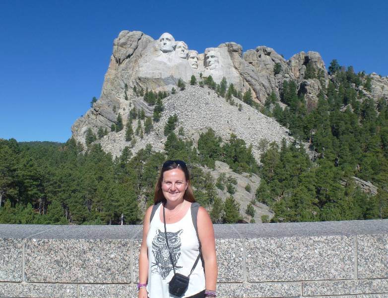 A smiling woman stands at the Grand View Terrace at Mount Rushmore National Memorial in South Dakota, with the granite carvings of George Washington, Thomas Jefferson, Theodore Roosevelt, and Abraham Lincoln visible in the background, framed by lush green pine trees under a clear blue sky.