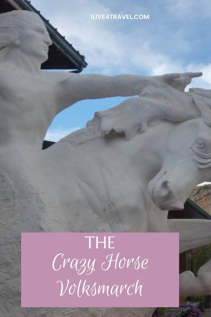 Pinterest pin image featuring a close-up of the white model statue of Crazy Horse pointing forward while riding a horse, with a partly cloudy sky in the background. The text on the pin reads: "The Crazy Horse Volksmarch"
