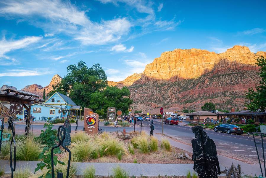 A colorful small-town street scene near Zion National Park at sunset, with warm light illuminating the towering red cliffs in the background. The foreground features Oscar’s Café sign, outdoor sculptures, and charming local buildings surrounded by greenery, while tourists and cars add a lively atmosphere.