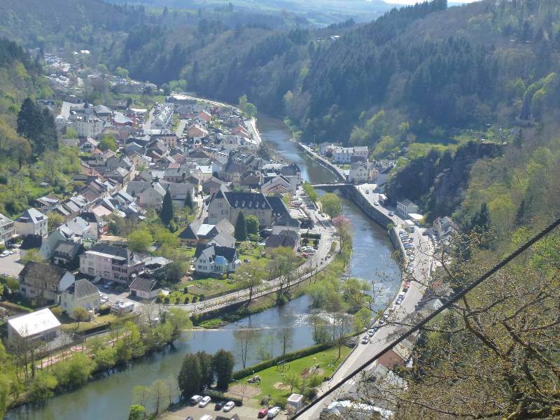 a river running through a valley, with a road to the right of it and houses on the land to the left, with a hill with trees to the right of the river