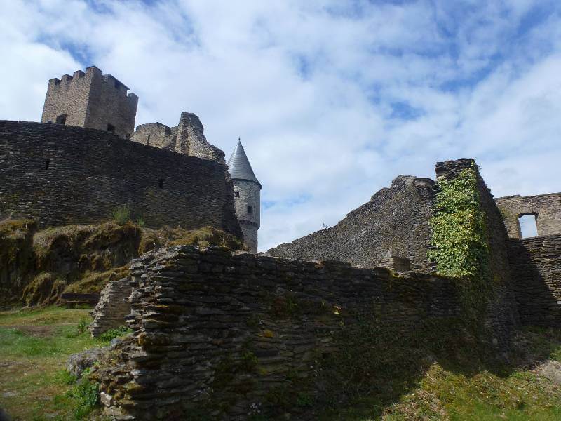 ruin walls of a castle, with a round turret with a roof on in the background. Grass on the floor and ivy covering part of the wall