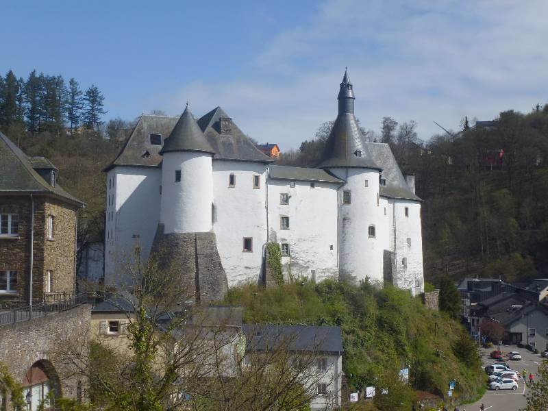 a white castle perched slightly higher than a town, with buildings to the left and below and a road to the right with trees behind it