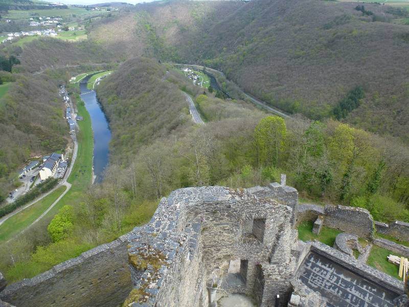 a river running like a horseshoe in a valley around the ruins of a castle, around the river we can see a few houses and the area is surrounded by hills with trees