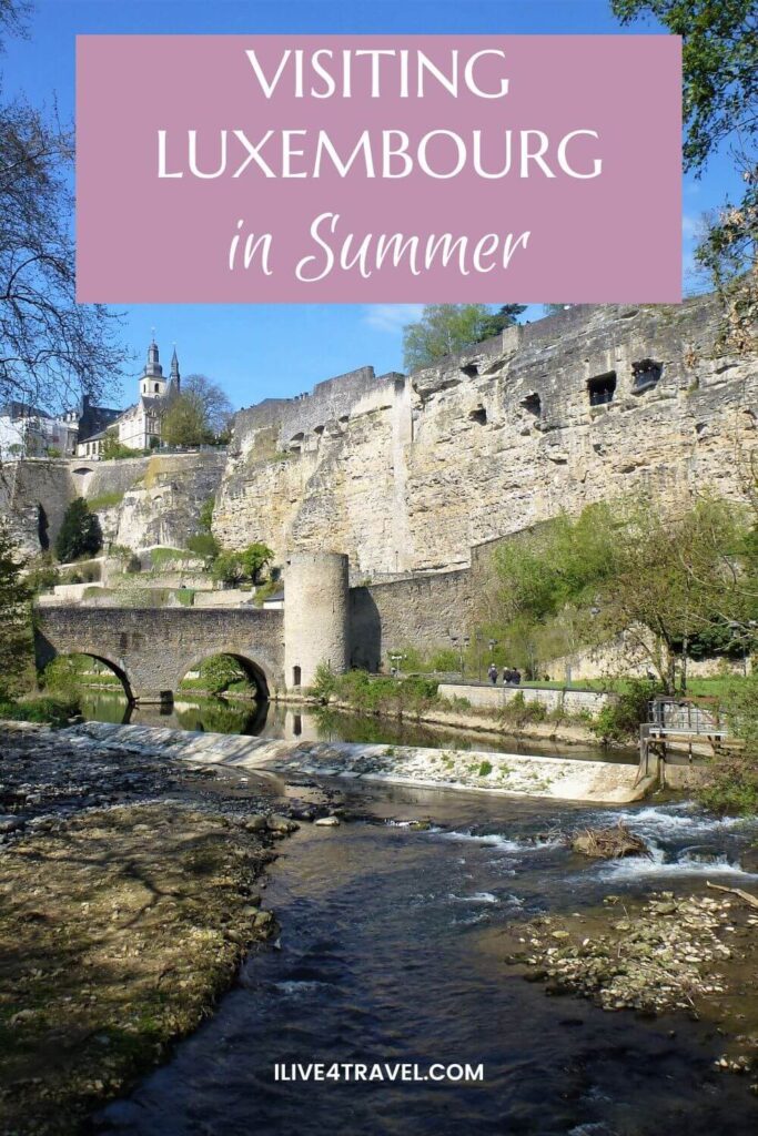 a pinterest pin about visitng luxembourg in summer, showing a river running through a valley and the valley wall has holes in the wall and an old stone bridge crossing the river on a sunny day