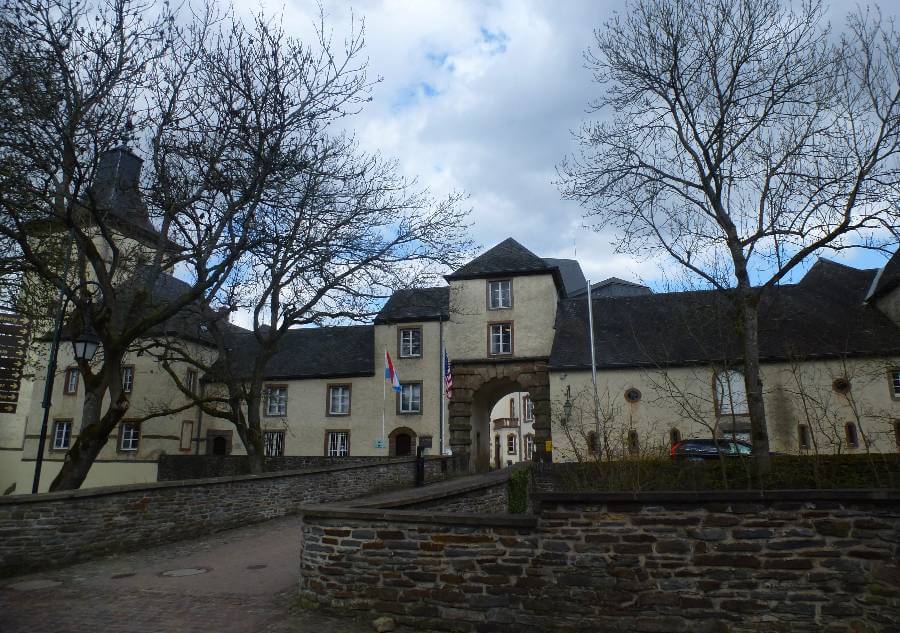 A view of a historic stone building complex in Luxembourg with an arched entranceway and flags displayed above it, including the Luxembourg and American flags. The cream-colored buildings feature slate roofs and small windows, surrounded by bare trees and stone walls along a cobblestone path under a partly cloudy sky.