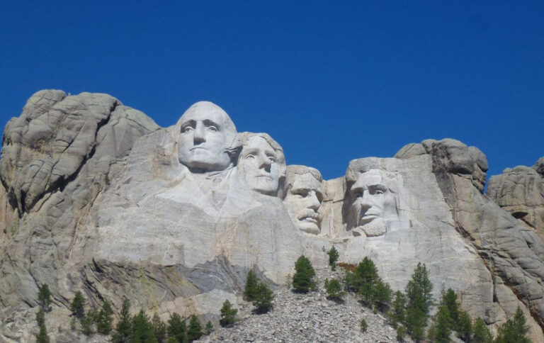 A clear daytime image of Mount Rushmore National Memorial in South Dakota, featuring the massive granite carvings of four U.S. Presidents, George Washington, Thomas Jefferson, Theodore Roosevelt, and Abraham Lincoln, set against a deep blue sky, with pine trees visible at the base of the mountain.